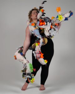 Image Description: A bird girl spreads her tangled wings, which are made from a patchwork of colourful fabrics, dangly string, and plastic toys.