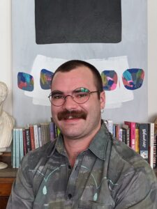 A portrait of Luke Maddaford smiling man with short brown hair and mustache. He is wearing glasses, and a grey patterned shirt. He is sitting in front of a row of books with a painting in the background.
