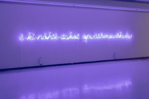 White empty room with a large glowing neon sign that says "ē-kī-nōhtē-itakot opwātisimowiskwēw" .