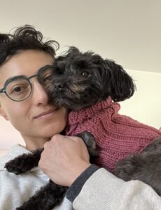 A person wearing a white hoodie and glasses holds a small black dog wearing a knitted sweater.