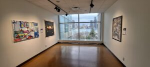 View of artworks hung in the community display space of graduating BFA students. Artwork is on the left and right side of the room , the back wall is windows facing Riverside Drive. 