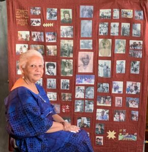 An older woman is pictured seated beside a quilt. She has short white hair and is wearing a blue dress.