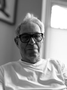 An older man pictured from the chest up, he has short white hair and wears black glasses with a thick frame. The image is in black and white.