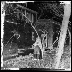 A photographic negative of a person wearing a dress, holding a basket and standing in front of a building.