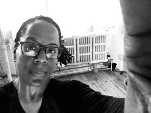 A black woman wearing glasses looks into the camera, she is visible in the lower left side of the image, and her hand, holding the camera is visible on the right. In the background is a bright room with wooden floors, large windows and heating registers.