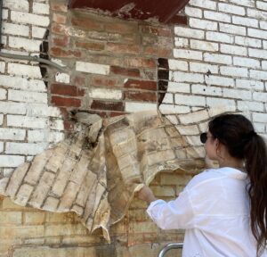 A woman with long dark hair in a ponytail, wearing a white collared shirt and black sunglasses pulls a latex cast from a brick wall.