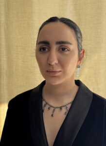 A woman staring straight ahead with her hair back in a bun, wearing a silver necklace and black blazer.