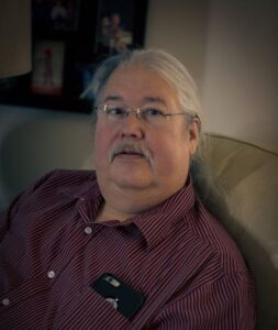 Russell Diabo is photographed sitting, his long grey white hair is pulled back and he is wearing a purple shirt and has a phone in his pocket, he is facing the camera.