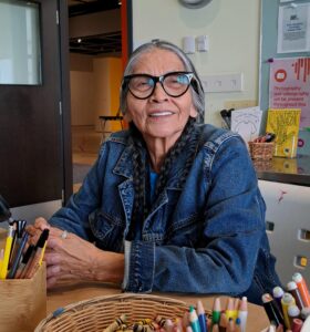 Dr Elder Mona Stonefish sits facing the camera smiling with her hands clasped. Her black and grey hair is braided, she is wearing black cat eye glasses and a denim jacket.