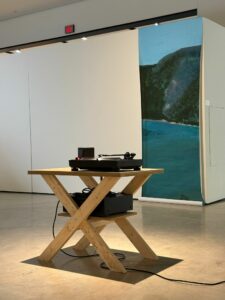 A record player sits on a wooden table in a gallery, with a tall seascape painting behind it.